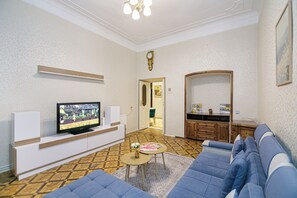 Een woonkamer met een televisie op een witte standaard, een blauwe bank, een klein rond tafeltje met een vaas en een vloer met een patroon.