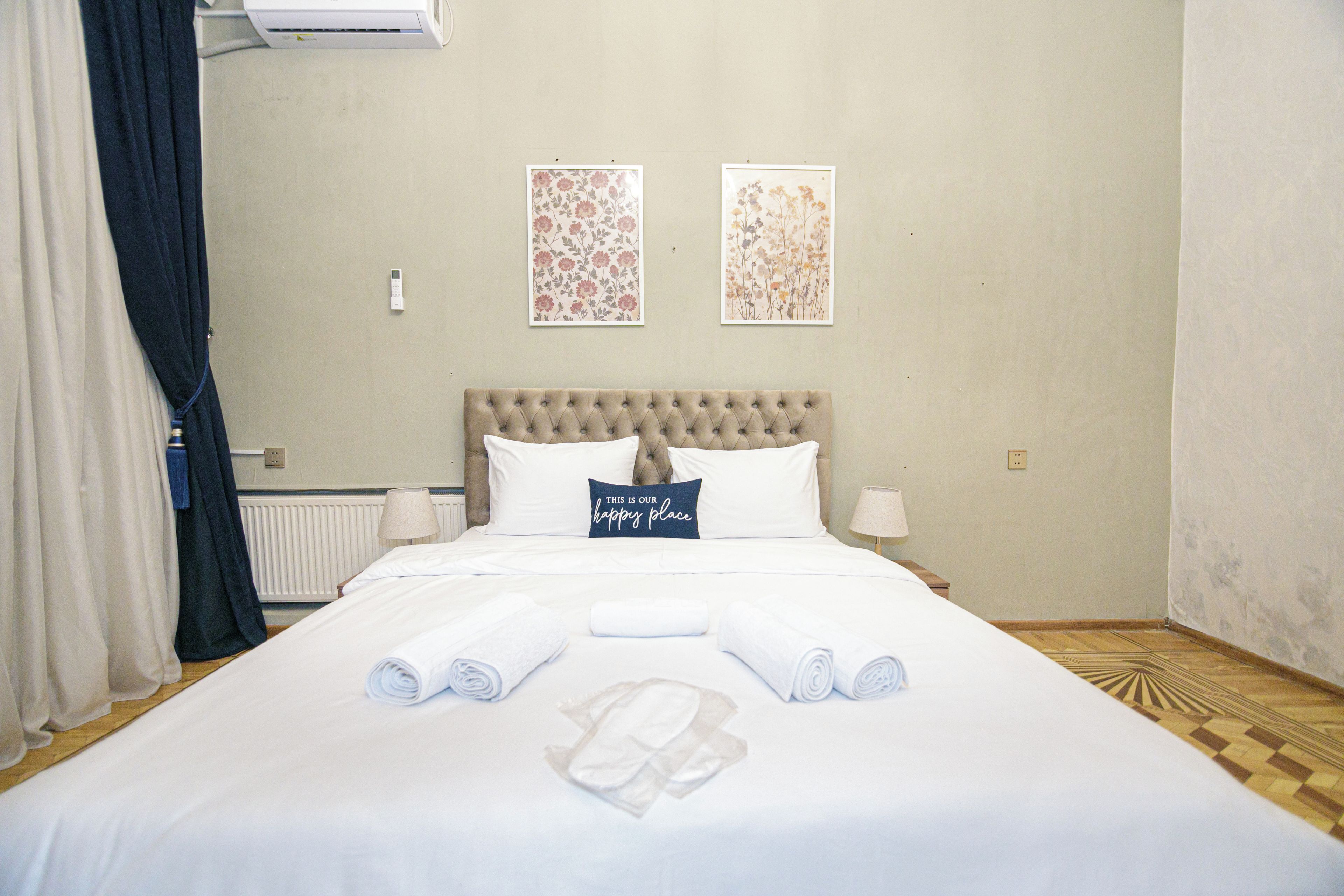 Una cama bien tendida con toallas enrolladas y un cojín con texto, una lámpara de noche y dos cuadros florales en la pared.
