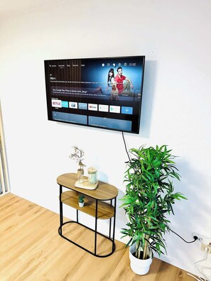 Un televisor de pantalla plana montado en la pared mostrando una película con subtítulos, una mesita de madera con una planta en maceta y un enchufe.