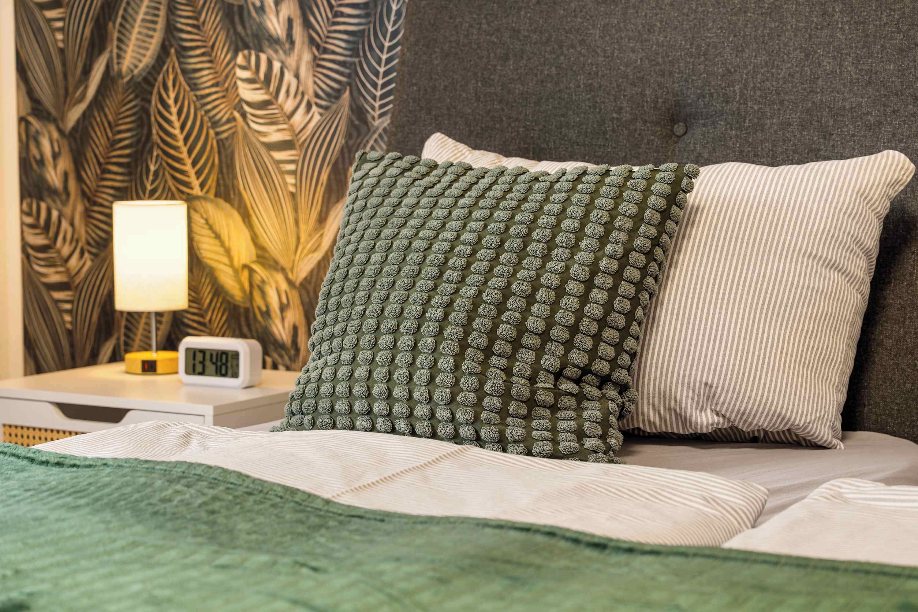 A neatly made bed with a green and white pillow, a bedside table with a lamp, and a wall with a leaf pattern.