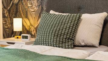 A neatly made bed with a green and white pillow, a bedside table with a lamp, and a wall with a leaf pattern.