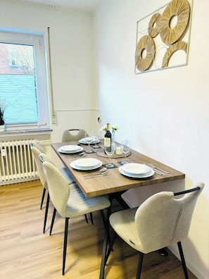 Un comedor con una mesa de madera dispuesta para cuatro personas, sillas, un radiador, una ventana con persianas y un adorno en la pared.