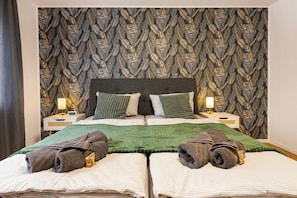 Une chambre d’hôtel avec un papier peint à motifs de feuilles, un lit avec une literie verte et blanche, deux oreillers et deux tables de chevet avec des lampes.