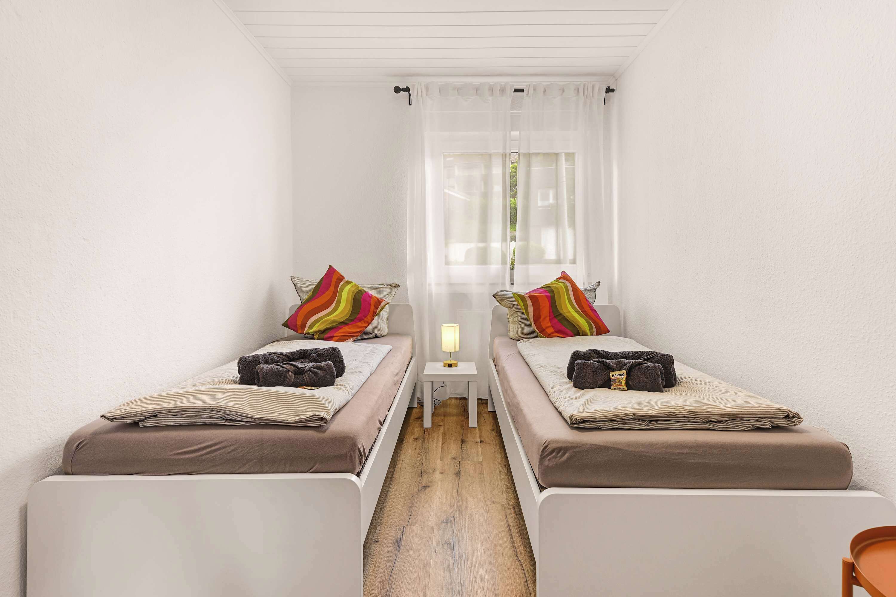 Een nette, kleine kamer met twee bedden, een nachtkastje en een raam met dunne gordijnen.