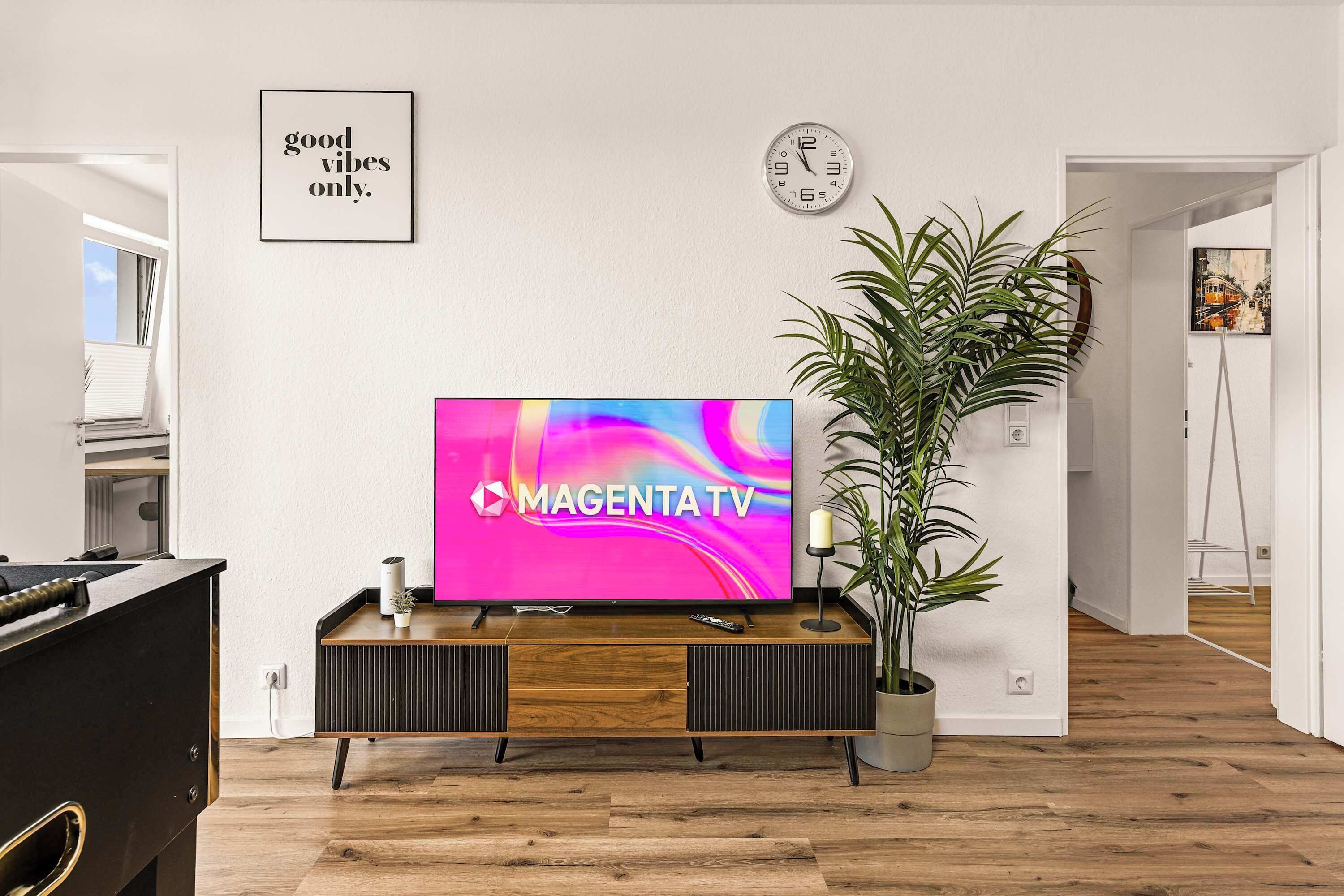 Ett modernt vardagsrum med en platt-TV, en möbel i trä för underhållningssystemet, en krukväxt och en väggklocka.
