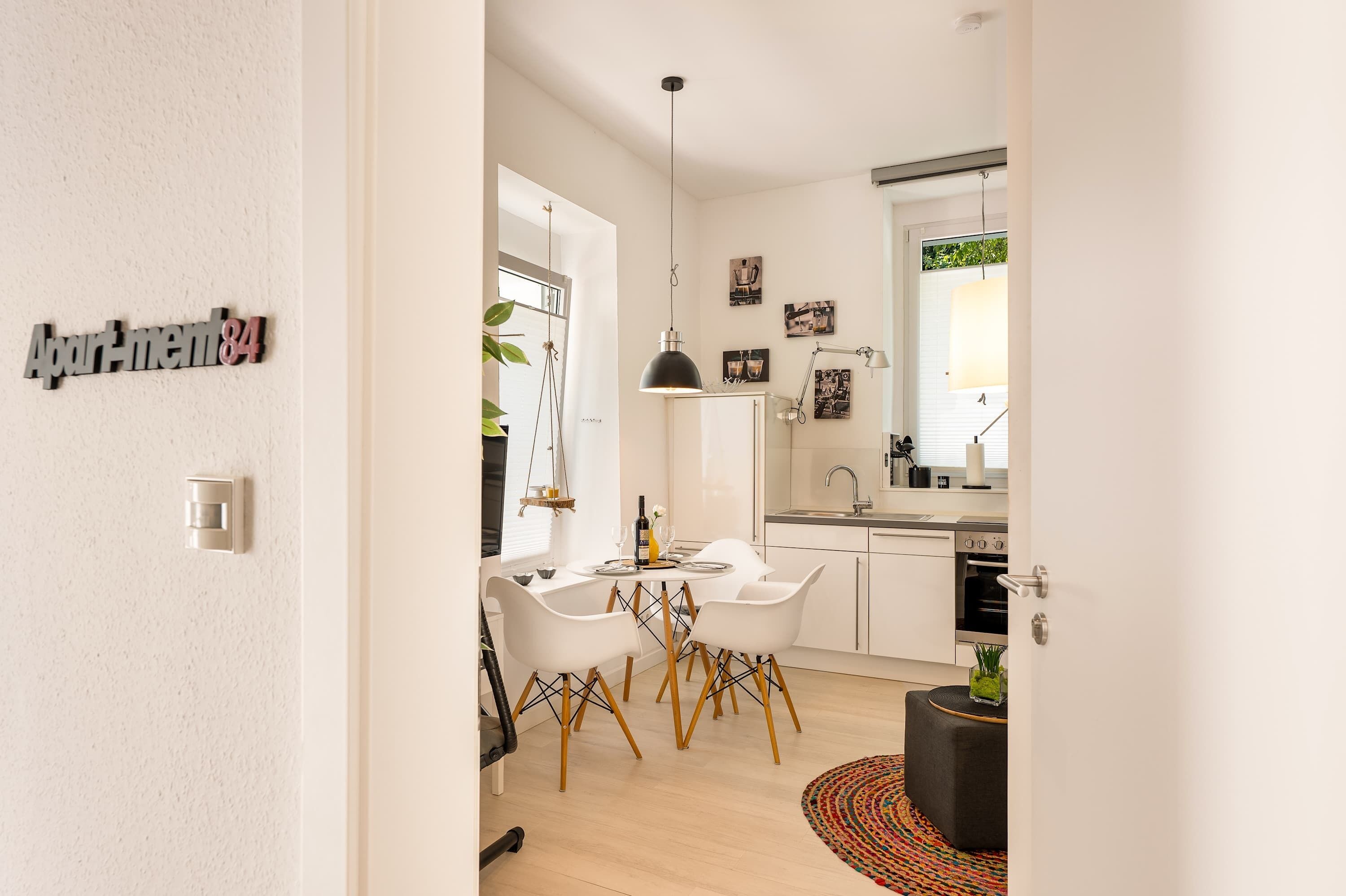 Eine moderne Küche mit weißen Schränken, einer schwarzen Pendelleuchte und einem Essbereich mit weißen Stühlen und einem Holztisch.