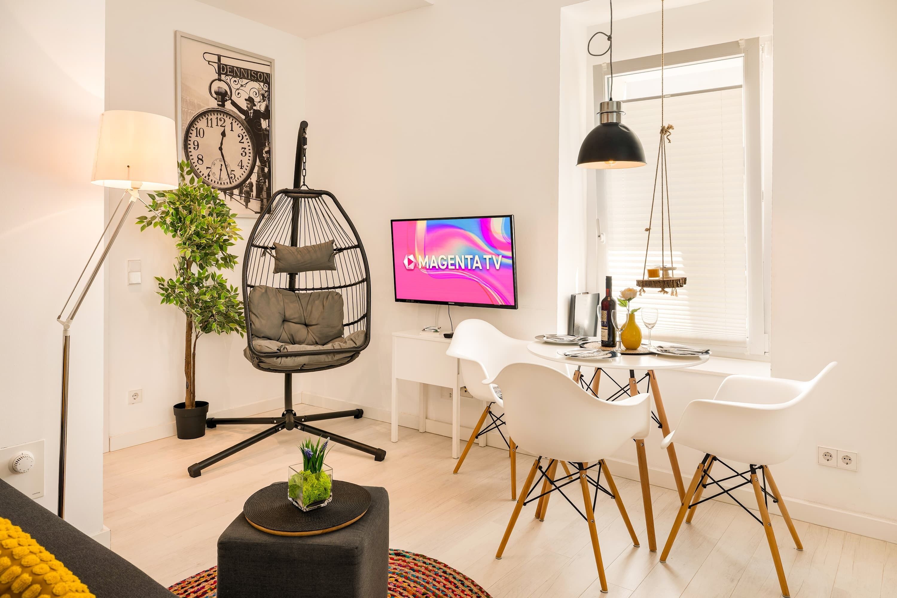 A modern living room with a hanging chair, a dining area with white chairs, a flat-screen TV, and a large window.