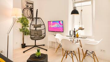 A modern living room with a hanging chair, a dining area with white chairs, a flat-screen TV, and a large window.
