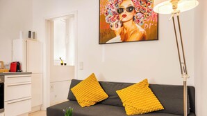 A modern living room with a grey sofa, two yellow cushions, a glass vase with green plants, and a painting of a woman with colorful flowers on the wall.