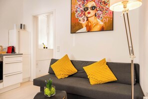 A modern living room with a grey sofa, two yellow cushions, a glass vase with green plants, and a painting of a woman with colorful flowers on the wall.