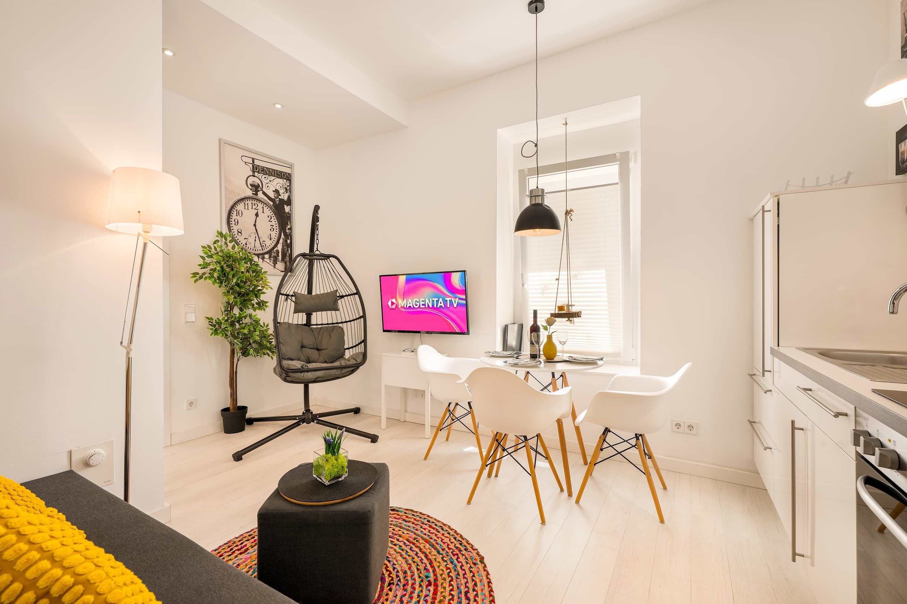 A modern living room with a sofa, a hanging chair, a dining table with chairs, a television, and a kitchen area with a sink.