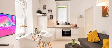Una cocina moderna con armarios blancos, un comedor con mesa y sillas blancas, y un sofá gris con cojines amarillos.