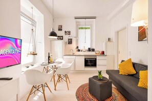 Une cuisine moderne avec des meubles blancs, un coin repas comprenant une table et des chaises blanches, et un canapé gris agrémenté de coussins jaunes.