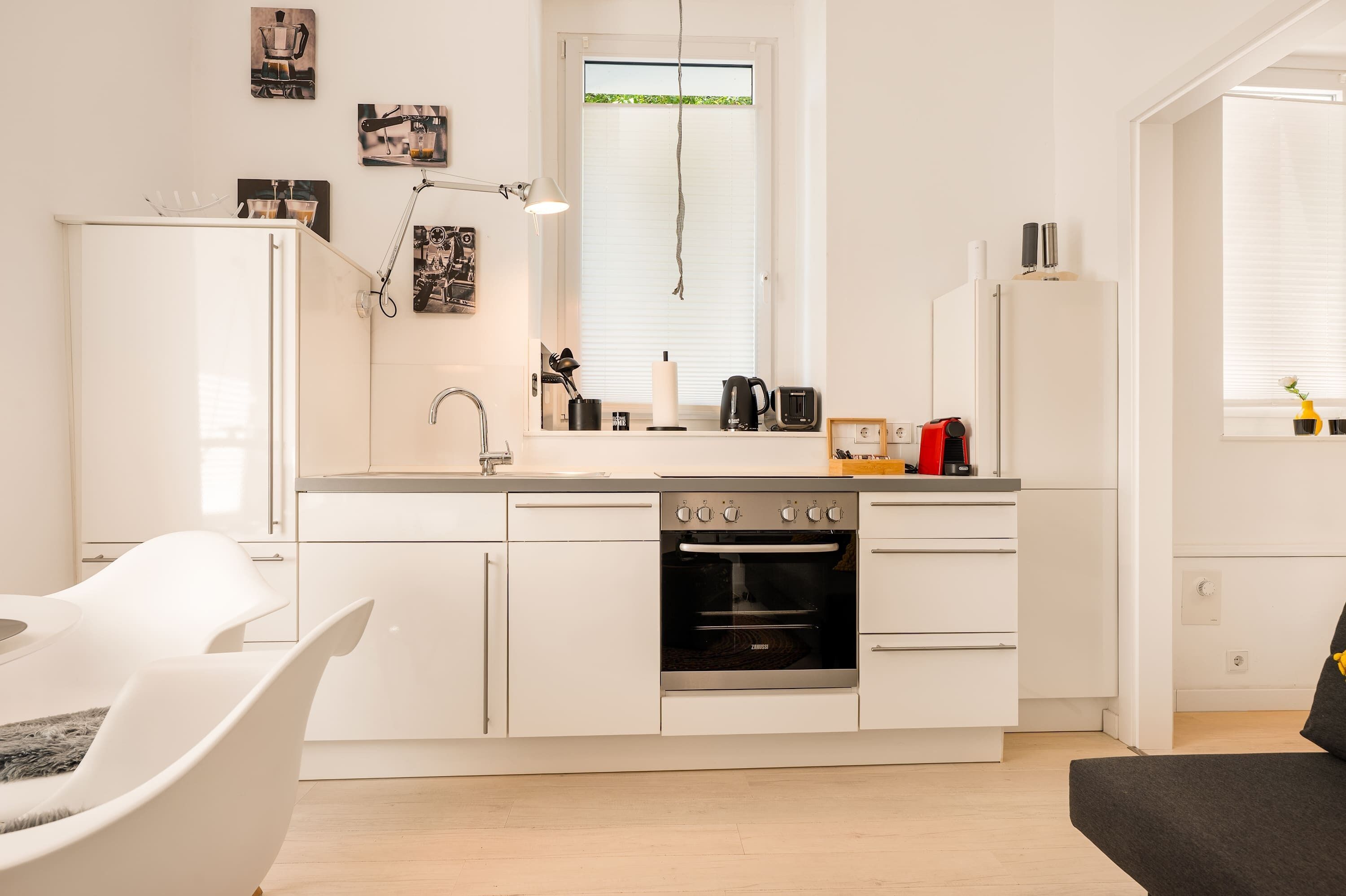Eine moderne Küche mit weißen Schränken, einem eingebauten Backofen, einer Kaffeemaschine und einem Fenster mit Jalousien.