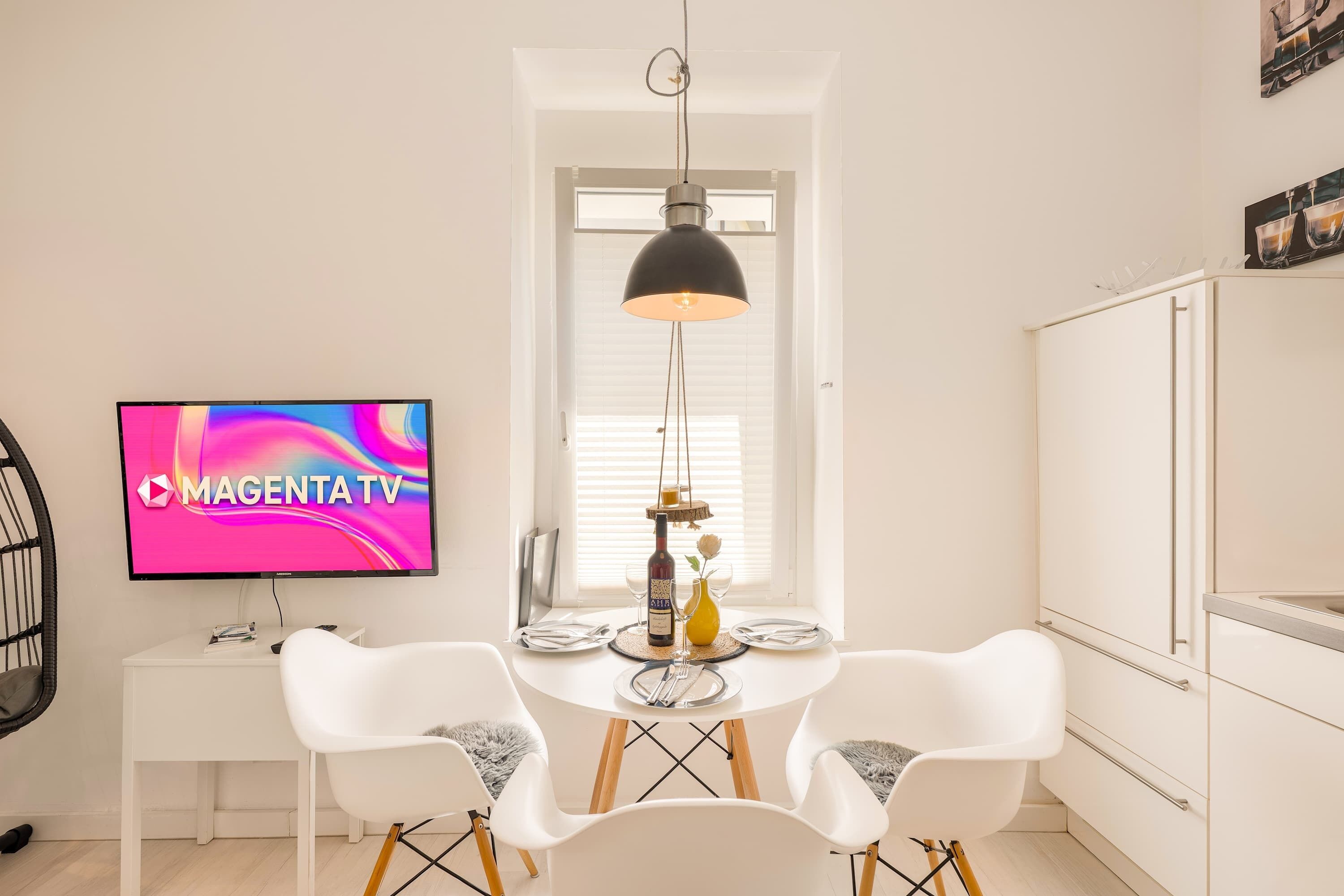 Ein modernes Wohnzimmer mit Flachbildfernseher, einem Esstisch mit Stühlen, einer Pendelleuchte und einem Fenster mit Jalousien.
