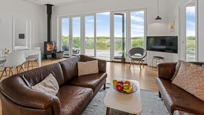 Amplia sala de estar con un sofá de cuero marrón, un televisor de pantalla plana, una chimenea y un gran ventanal corredero que da a una terraza con vistas al paisaje.