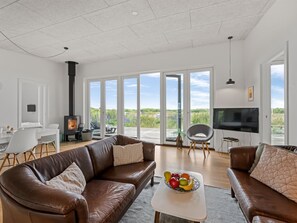 Un salon spacieux avec un canapé en cuir marron, un téléviseur à écran plat, une cheminée et une grande porte-fenêtre coulissante donnant sur une terrasse avec vue sur le paysage.
