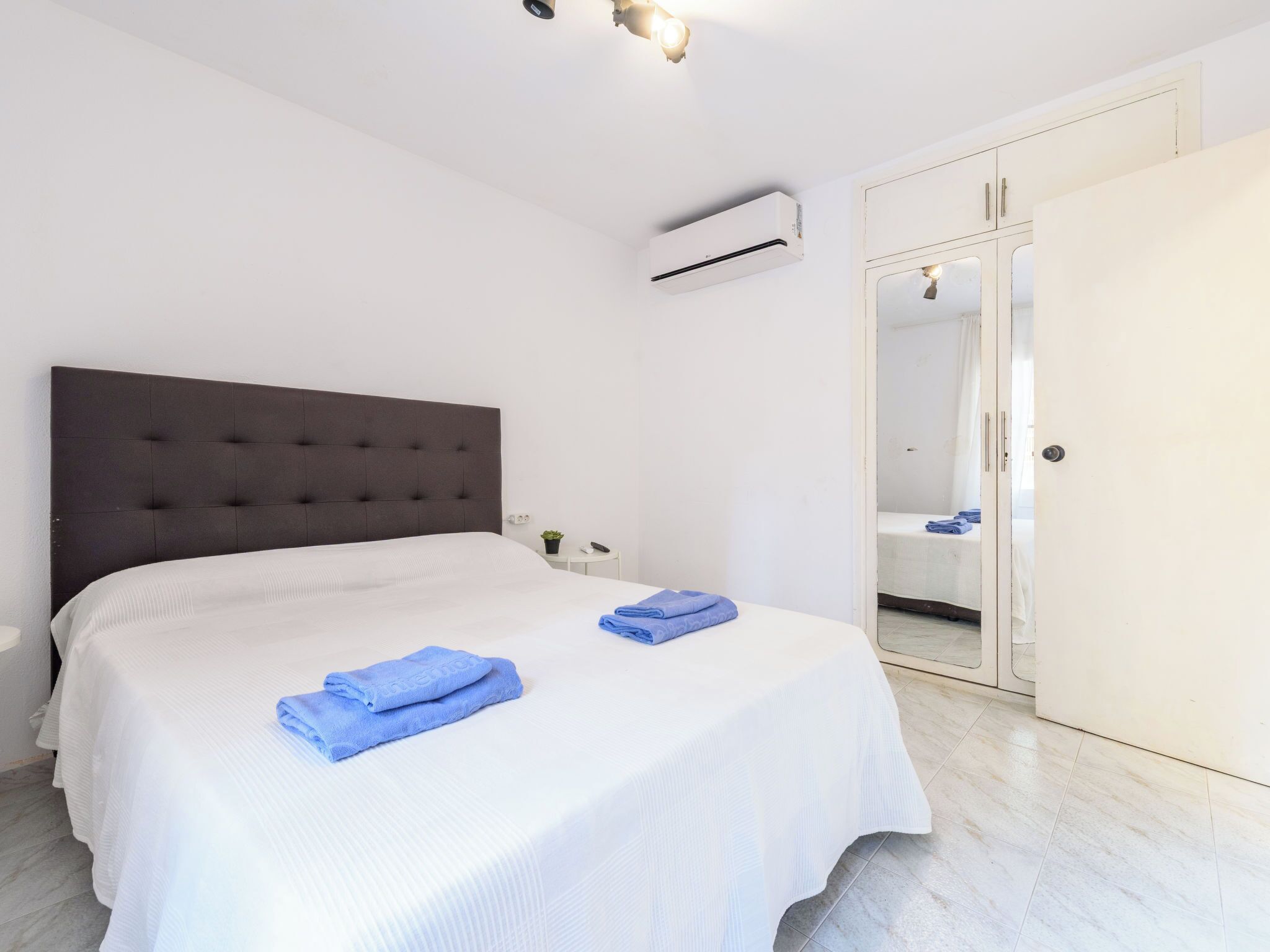 Un dormitorio con una cama grande, una mesita de noche, un equipo de aire acondicionado, un armario y un baño visible a través de una puerta de cristal.
