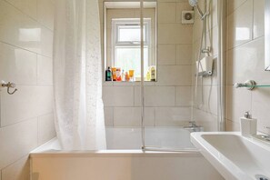 A bathroom with a bathtub, shower, and a window with toiletries on the windowsill. - Home Away From Home in Selhurst Park (London)