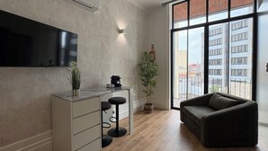 A modern living space with a flat-screen TV mounted on the wall, a small kitchen area with a coffee machine, and a large window offering a view of buildings. - Passos Collection 263 (Porto)