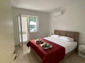 A hotel room with a bed, a window with blinds, a door, and a white air conditioning unit. - Two bedroom apartment with garden (Trpanj)