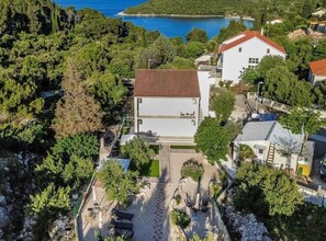 An aerial view of a residential area with a white building featuring a red roof, surrounded by greenery and a body of water in the background. - Two bedroom apartment with garden (Trpanj)