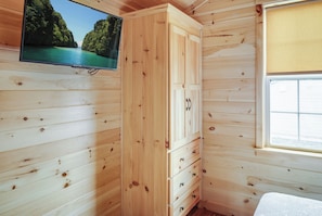 A wooden cabin interior with a television mounted on the wall, a window with blinds, and a bed with a white bedspread. - Jellystone Park Gardiner (Gardiner)