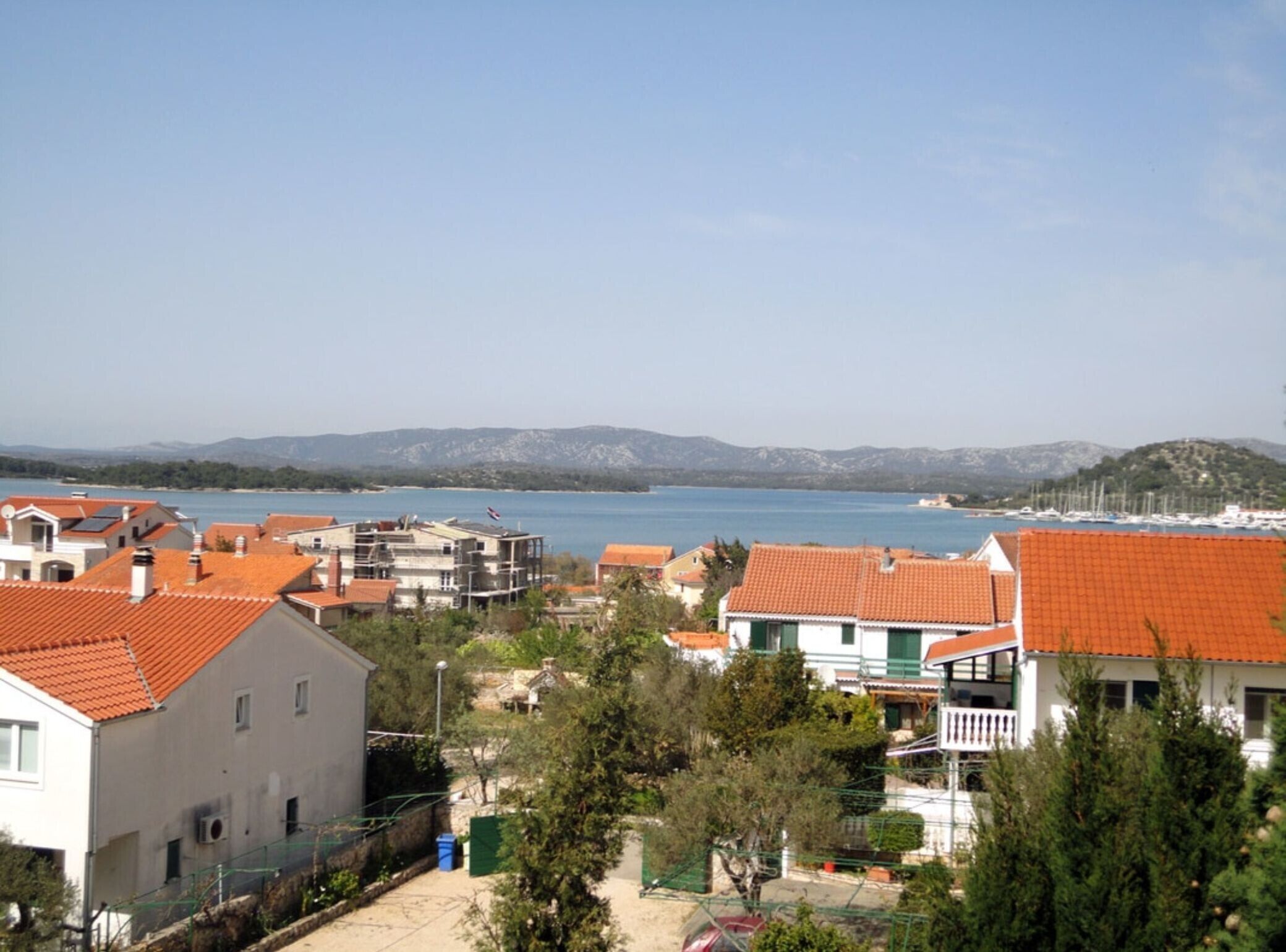 A coastal residential area with terracotta-roofed houses, a marina, and a clear view of the sea and distant mountains.