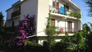 Ein zweistöckiges Gebäude mit Balkonen, einem sichtbaren Fenster und einem Baum mit violetten Blüten.