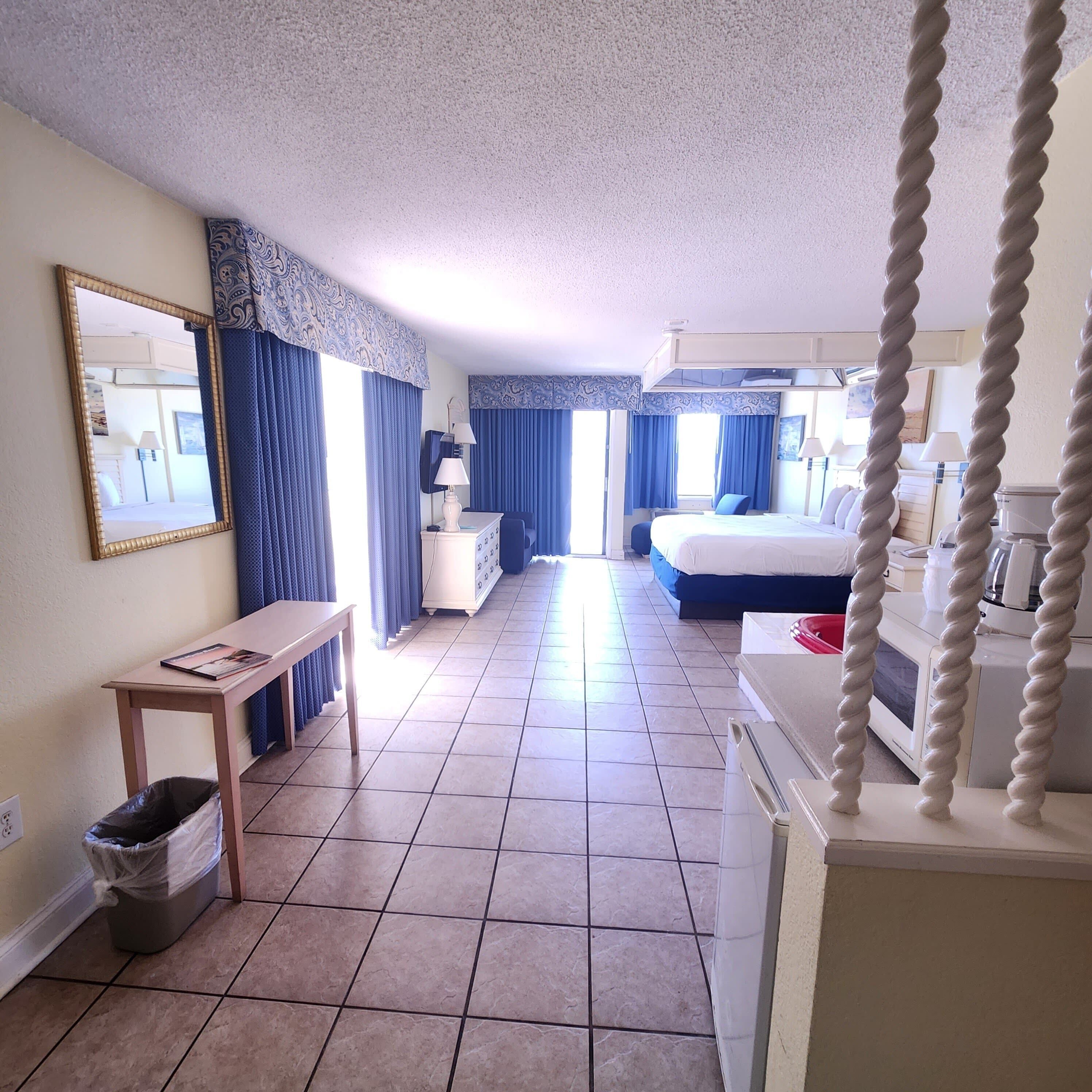 A hotel room with a bed, a desk, a chair, a trash bin, a mirror, and a television.