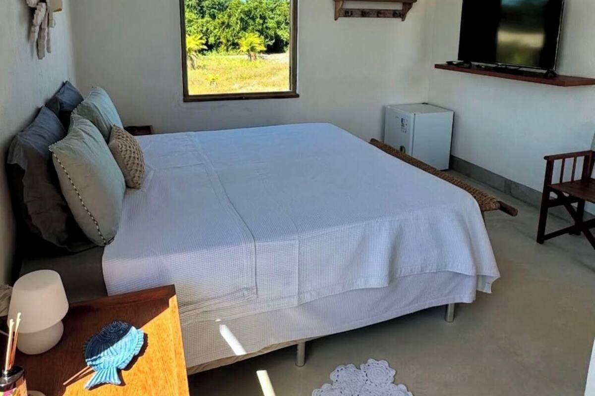 A bedroom with a large bed, a TV, a small fridge, a window with a view of greenery, and a wooden shelf with a plant.