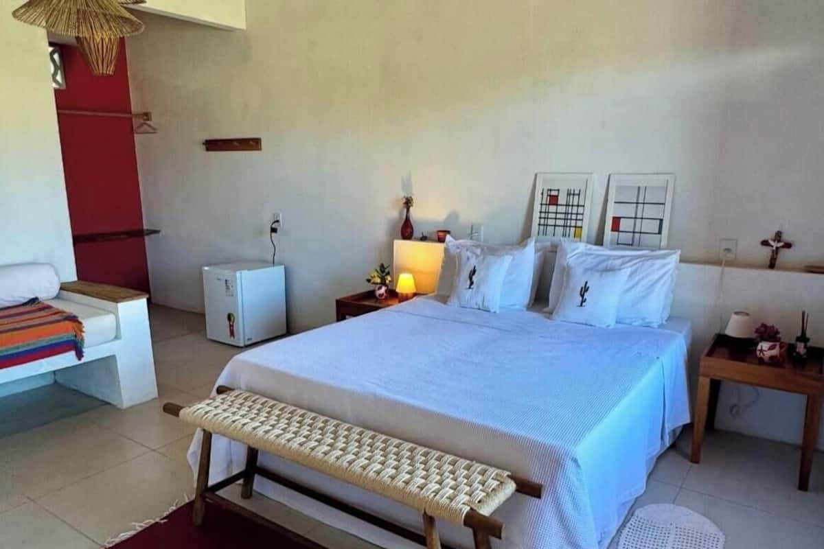 A bedroom with a bed, bedside tables, a bench, a hanging light fixture, and a small refrigerator.
