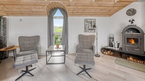 Ett mysigt rum med ett träpanelat tak, två grå fåtöljer, ett litet bord, ett fönster med gardiner, en väggklocka och en vedeldad kamin.