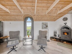 A cozy room with a wooden ceiling, two grey armchairs, a small table, a window with curtains, a wall clock, and a wood-burning stove. - "Janne" -  from the sea by Interhome (Ringkøbing)