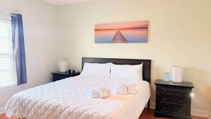 Een netjes opgemaakt bed met witte lakens, een donker hoofdbord en een schilderij van een zonsondergang boven het water aan de muur.