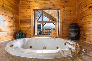 Ein Holzhütten-Innenraum mit einer großen, ovalen Badewanne, einem Fenster mit Ausblick und einer schwarzen Vase mit Adler-Motiv.