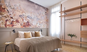 Ein Schlafzimmer mit einer großen Wandmalerei, einem Bett mit vielen Kissen, einem Nachttisch und einem Kleiderhaken mit Kleidung.