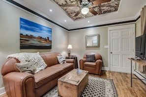 Un salon avec un canapé en cuir marron, une table basse en bois, une table d’appoint avec une lampe, un tableau encadré au mur et un ventilateur de plafond.