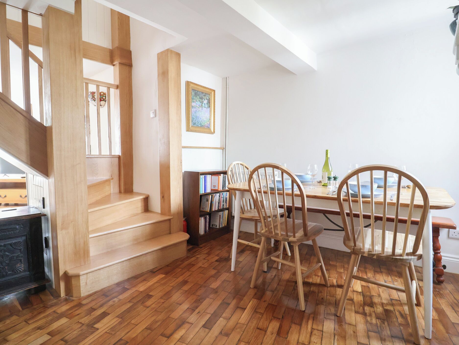 Un comedor con sillas y una mesa de madera, una escalera con barandillas de madera y una estantería llena de libros.