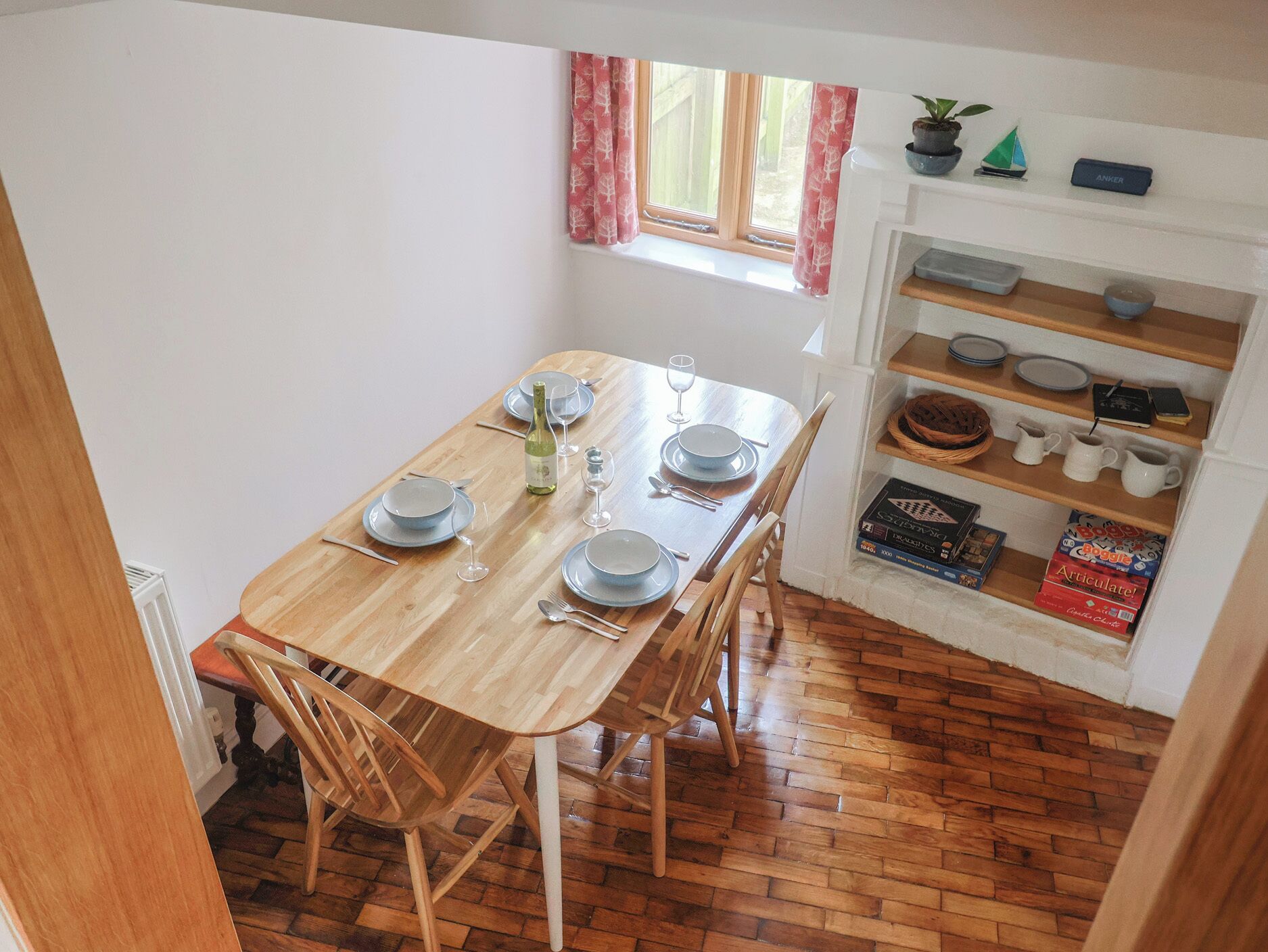A dining area with a wooden table set for four, a window with curtains, and a shelf with kitchen items.