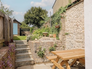 A stone wall with a wooden bench and steps leading to a garden area with plants and a blue building in the background. - Saybrook Cottage (Bridport)