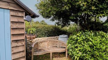 A wooden bench in a garden with a stone wall, a blue building, and a tree.