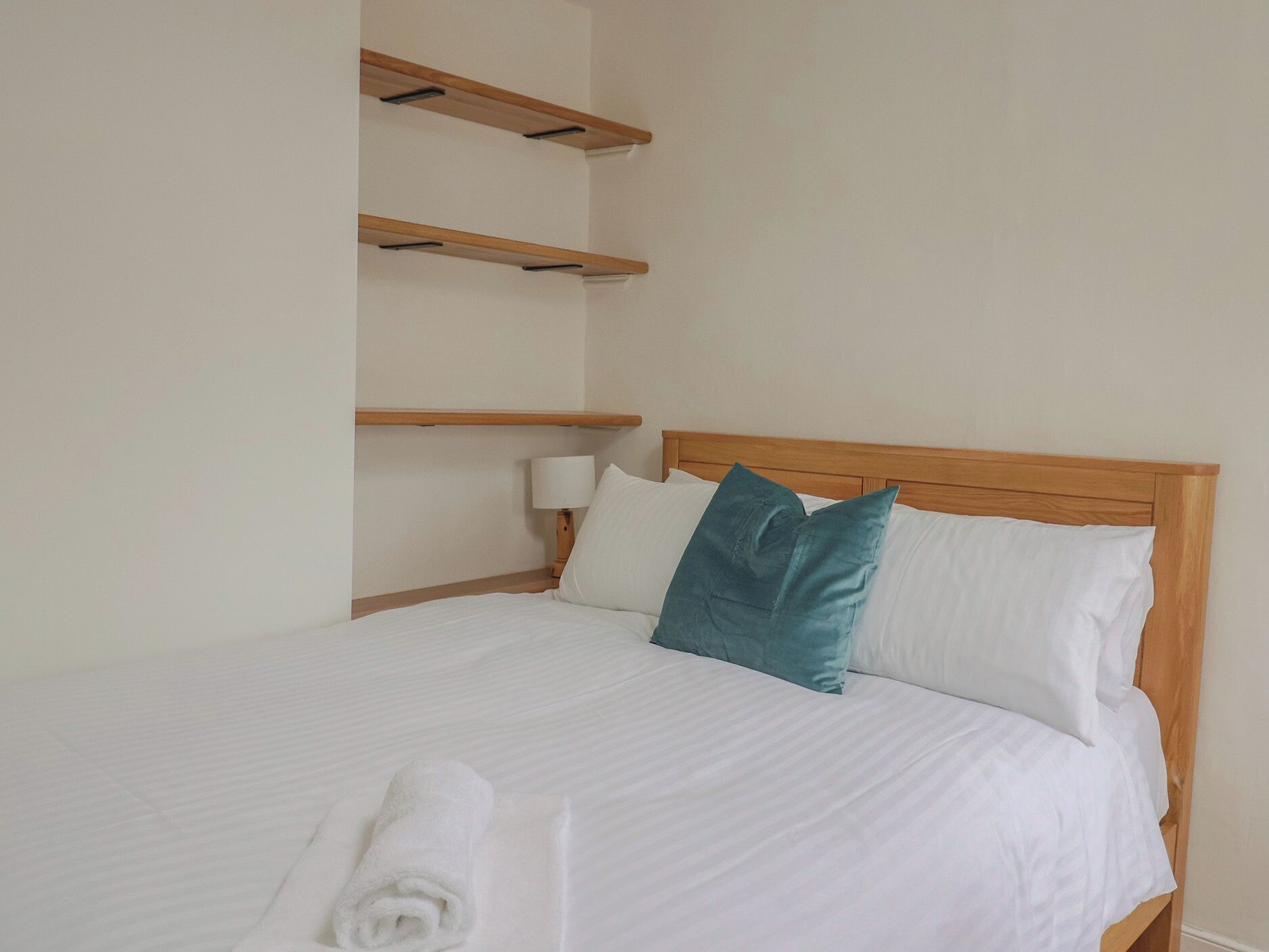Una cama bien hecha con ropa de cama blanca y una almohada verde azulada, un cabecero de madera y una pared blanca con un estante sobre la cama.