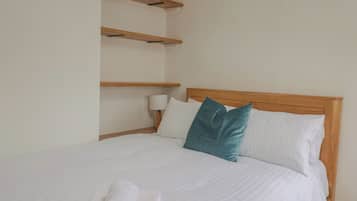 Un lit bien fait, avec une literie blanche et un coussin turquoise, une tête de lit en bois et un mur blanc sur lequel est fixée une étagère au-dessus du lit.