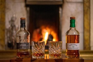 Three bottles of single malt Scotch whisky and two glasses on a wooden surface with a fireplace in the background. - Western Isles Hotel (Tobermory)