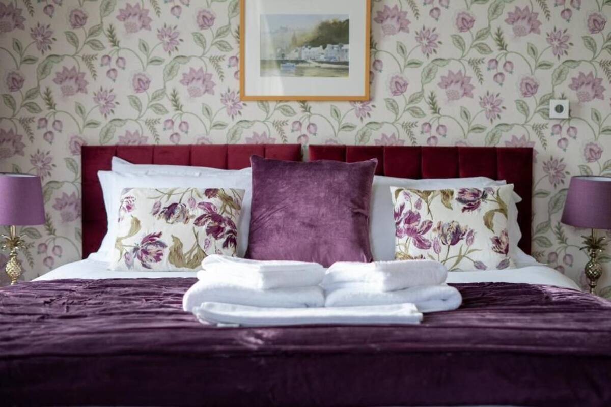 A neatly made bed with floral pillows and a folded towel, a red headboard, and a framed picture on the wall.
