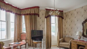 A room with a television on a stand, a chair, a desk with a lamp, and curtains with a floral pattern.