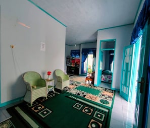 Una habitación con alfombra estampada, dos sillas, una mesita con un florero y una cama con cubrecama colorido.