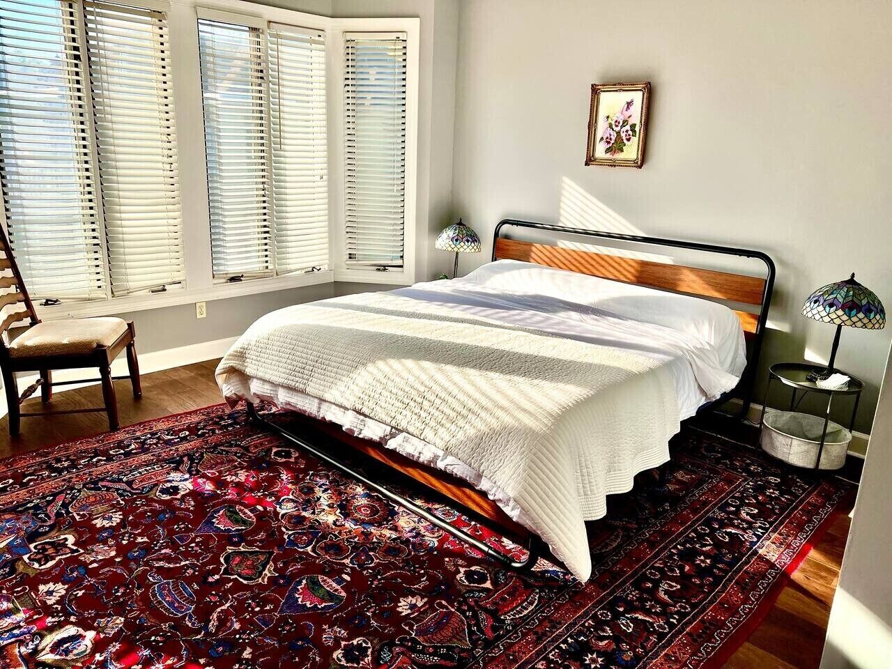 A bedroom with a bed, a chair, a nightstand, a lamp, and a window with blinds.