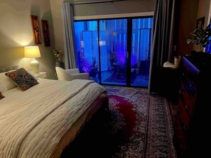 Ein Schlafzimmer mit Bett, Nachttisch, Lampe, Sessel und einem großen Fenster mit einer Schiebetür zu einem Außenbereich.